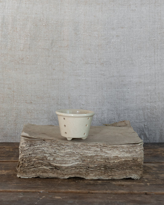 A small ceramic cup with a pale greenish-gray glaze and small perforations, displayed on a rustic wood block against a textured gray linen backdrop.