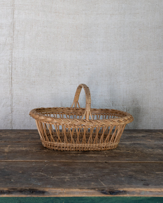 A natural woven wicker basket with a single handle, placed on a rustic dark wooden surface against a light gray textured wall.