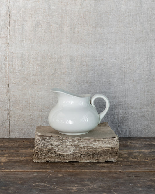 A glossy white ceramic pitcher with a curved spout and loop handle, displayed on a rustic wood block against a textured gray linen backdrop and dark wooden table.